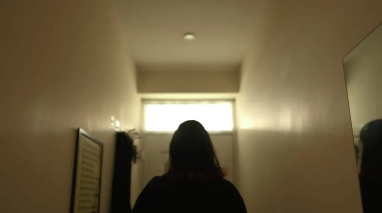 Person with long dark hair walking towards a bright doorway in a narrow, minimalist white corridor.
