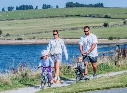 Two adults and two young children on a path beside a blue reservoir, with rolling green hills beyond.