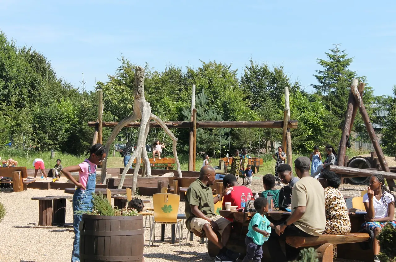 Families and children enjoy outdoor play equipment and seating in a woodland adventure area with climbing structures and wooden installations.