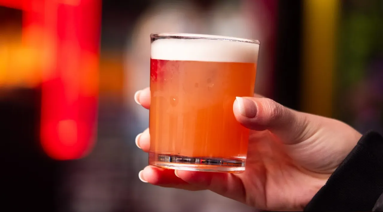 A hand holds a glass of amber beer with a white head against a blurred colourful background.