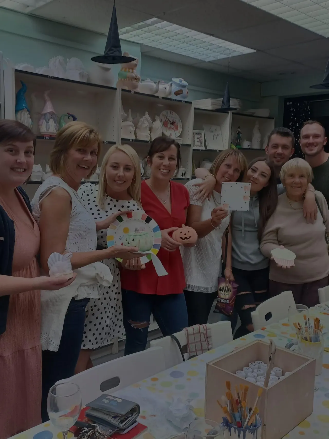 A group of adults posing together in a pottery studio decorated with wizard hats, displaying painted ceramic pieces and craft projects.