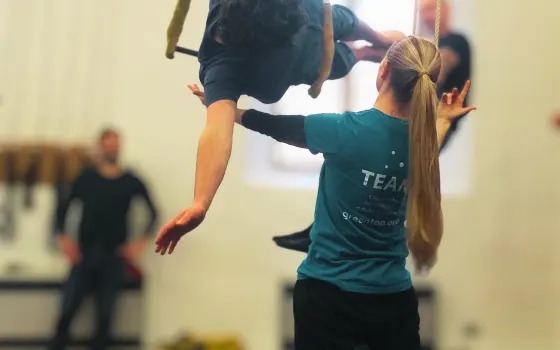 A child in a teal team shirt supports an adult performing an inverted acrobatic move indoors.