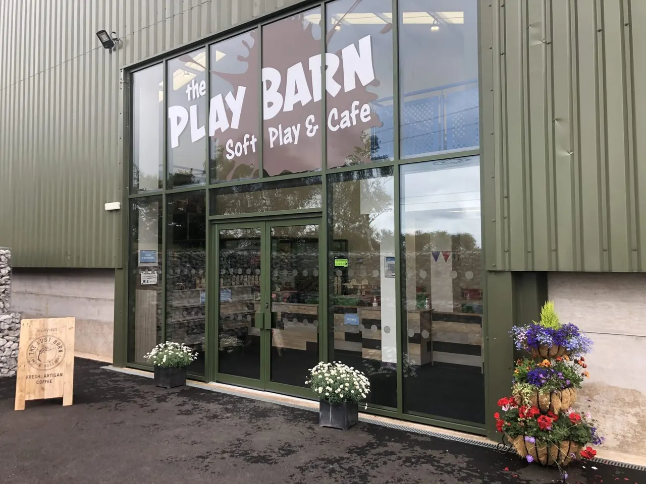 Exterior of a soft play venue with green-framed glass doors and windows, flower baskets outside, and a wooden coffee sign to the left.