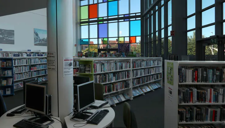 Library interior with bookshelves, computer workstation, and colourful stained glass windows overlooking outdoor scenery.