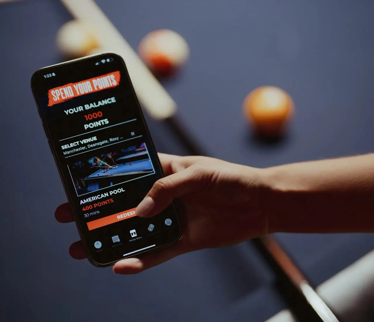 Hand holding a smartphone displaying a loyalty rewards app with points balance, set against a blurred background of pool balls and a cue.