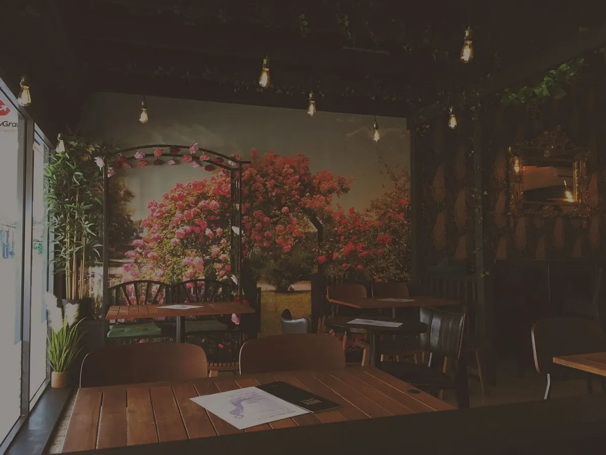 Evening view of a restaurant dining area with wooden tables, chairs, and a large mural of pink flowering vines and a garden bench on the back wall, lit by hanging pendant lights.