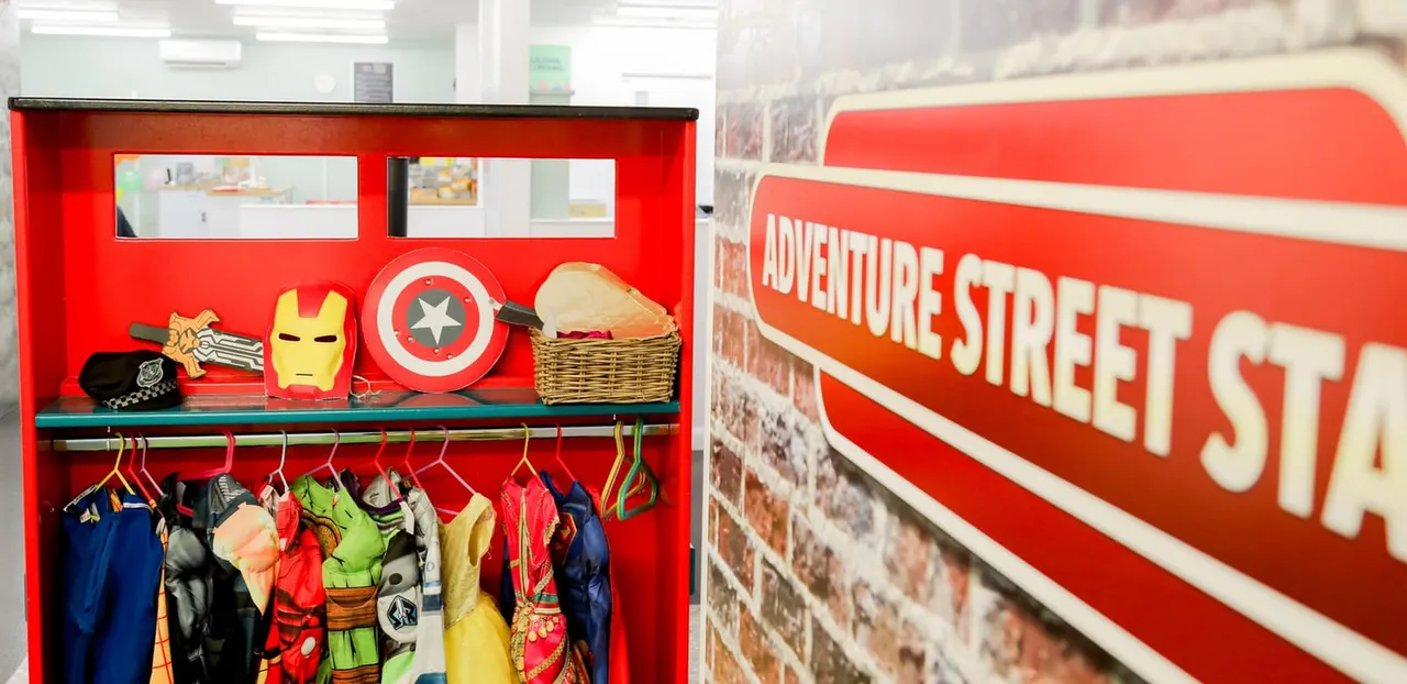 Red display unit with superhero-themed costumes, masks and shields hanging from rails, with red signage reading "Adventure Street" visible beside brick wall.
