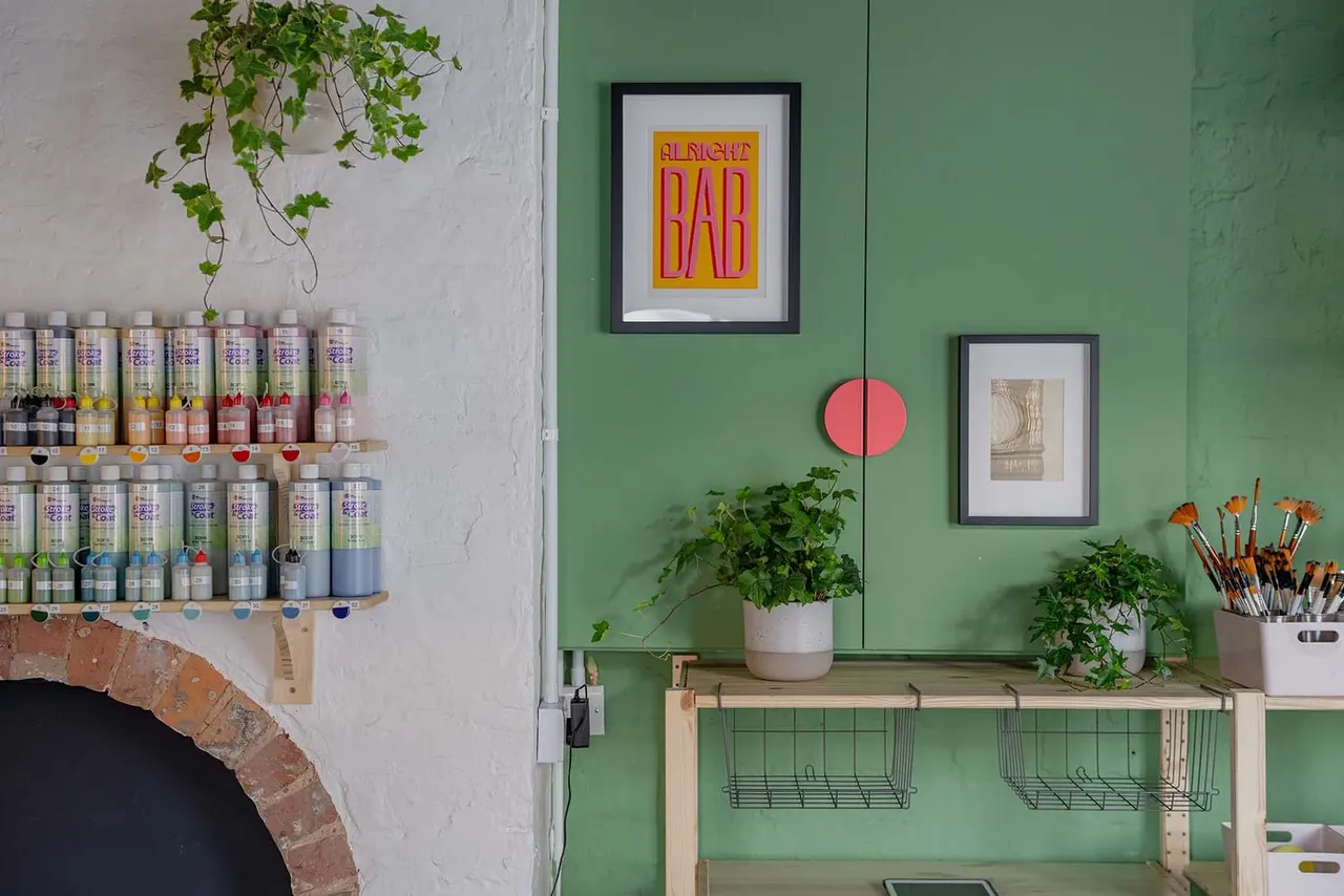 Art studio interior with shelves of paint bottles, framed signs on green wall, potted plants, and brushes in containers.