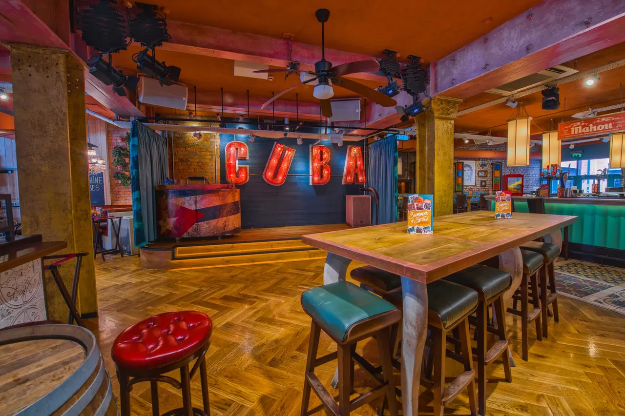 Colourful retro-themed bar interior with wooden flooring, high tables, stools, a stage with red neon "CUBA" sign, and vibrant orange and blue décor.