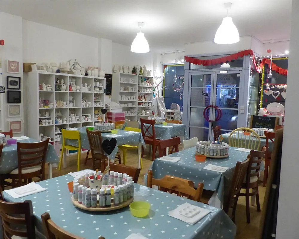 Brightly lit pottery studio with multiple tables set up for craft activities, shelves displaying finished ceramics, and festive red decorations.