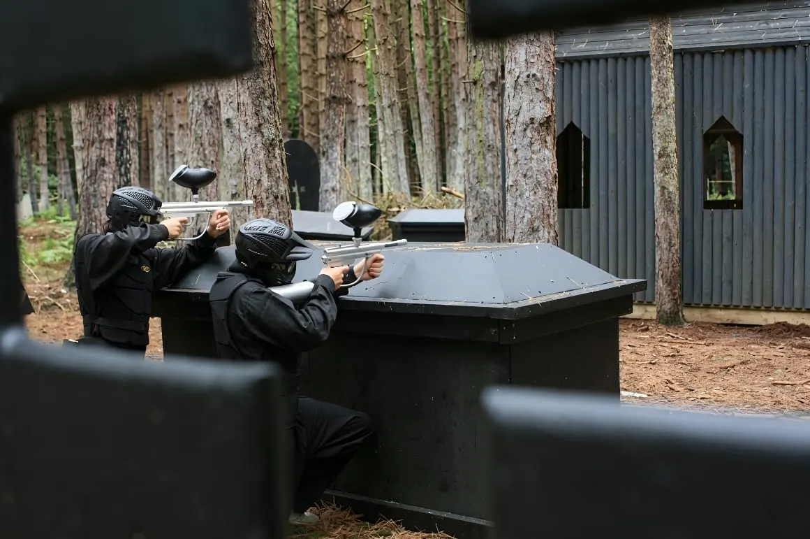 Players in protective gear and helmets playing paintball behind barriers in a wooded arena.