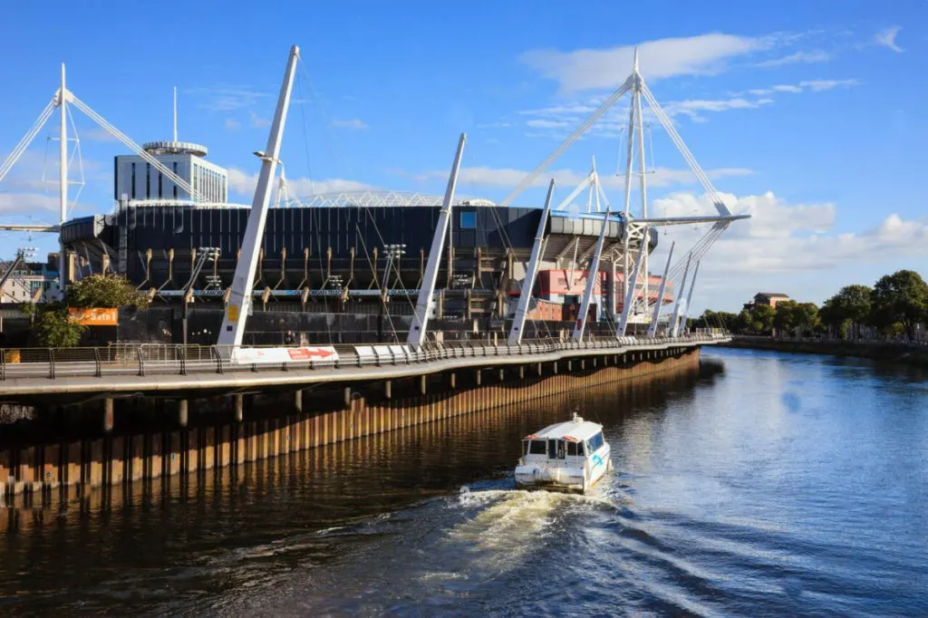 Modern riverside venue with distinctive white cable-stayed roof beside river, small white boat passing in foreground.