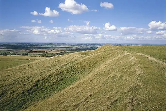 Rolling chalk downland with grass-covered Iron Age hill fort ridges overlooking distant farmland under blue sky.