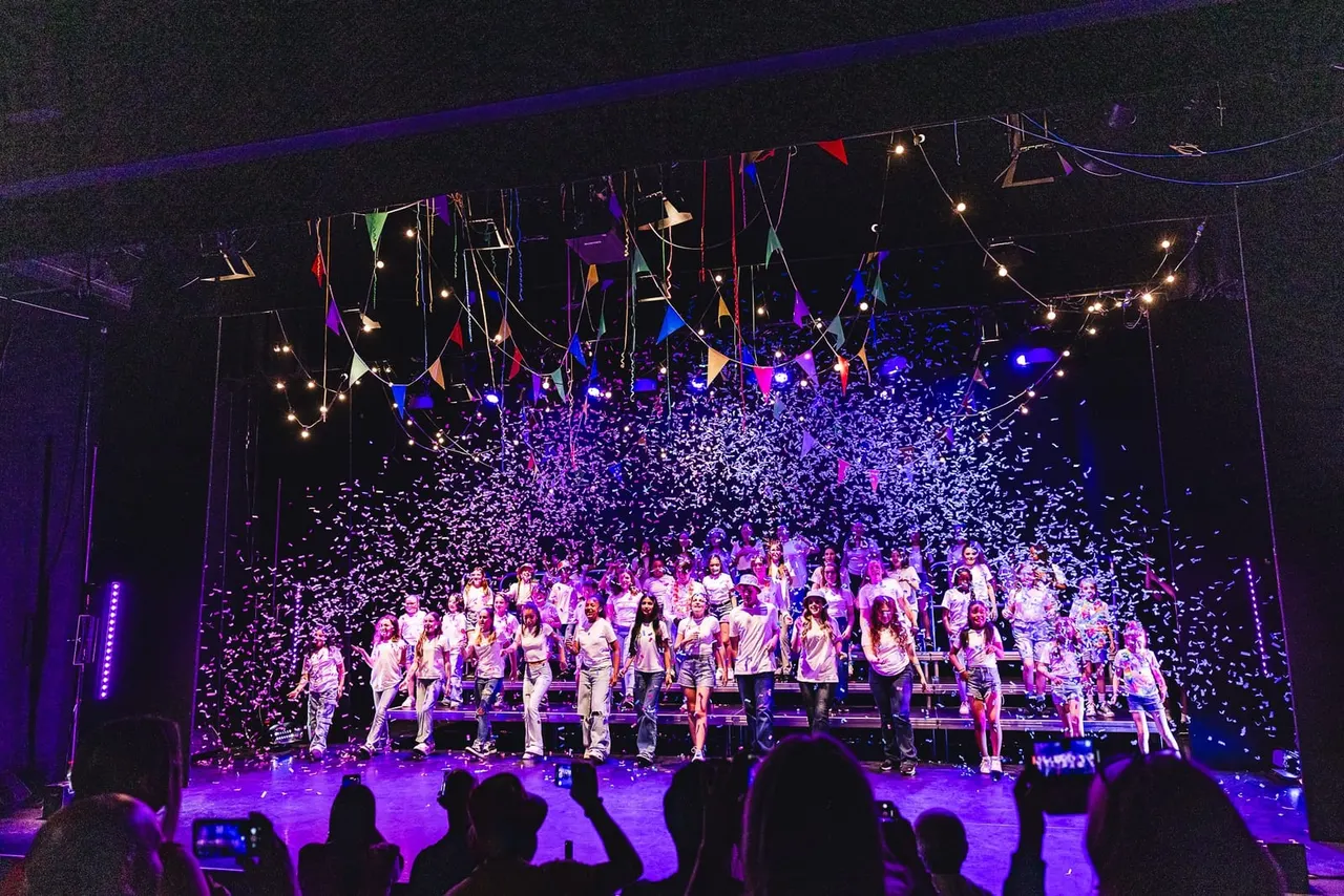 A large group of children and young people in white clothing perform on stage under purple lighting with confetti falling, whilst an audience watches from below.
