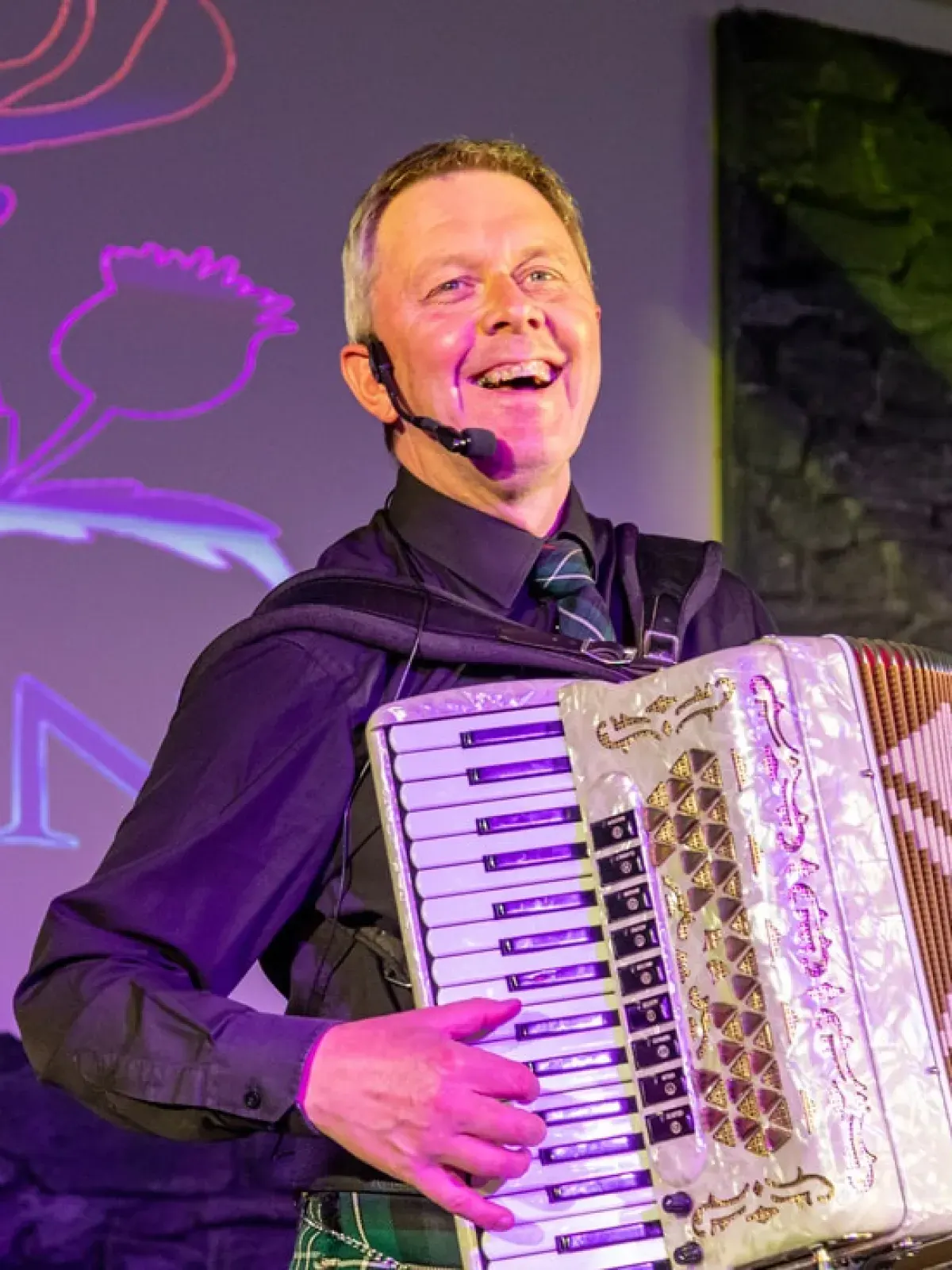 A man wearing a headset microphone performs on stage whilst holding an accordion, smiling broadly against a purple-lit backdrop.