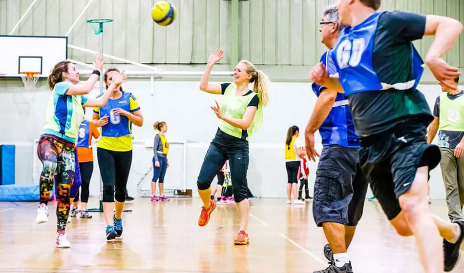 Mixed-age group playing volleyball indoors, with players jumping and passing a yellow ball in a sports hall.