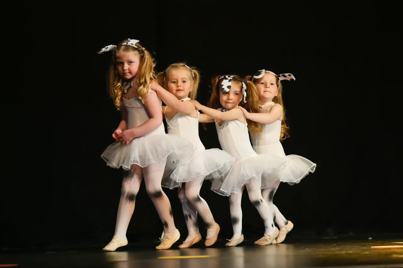 Four young ballet dancers in white tutus and cow-print headbands performing together on stage.