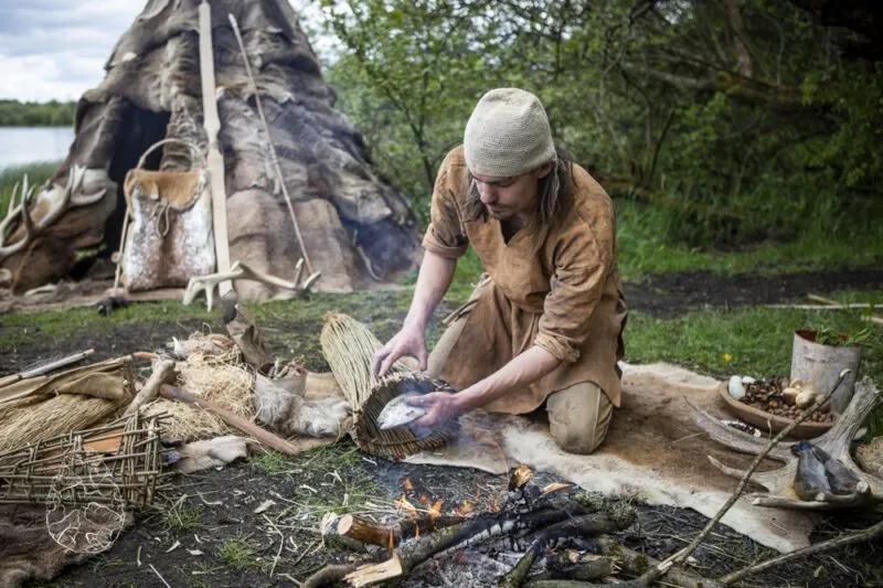 Person in period clothing preparing food at a campfire beside a traditional tipi shelter near water.