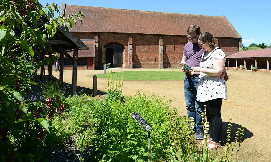 Two adults consult a map in a historic house courtyard garden with herb plantings and a brick Tudor building.