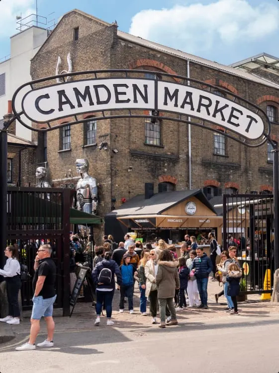 This is a PHOTOGRAPH.

Camden Market entrance with crowds of visitors gathered at the ornate metal sign gate in front of the historic brick building.