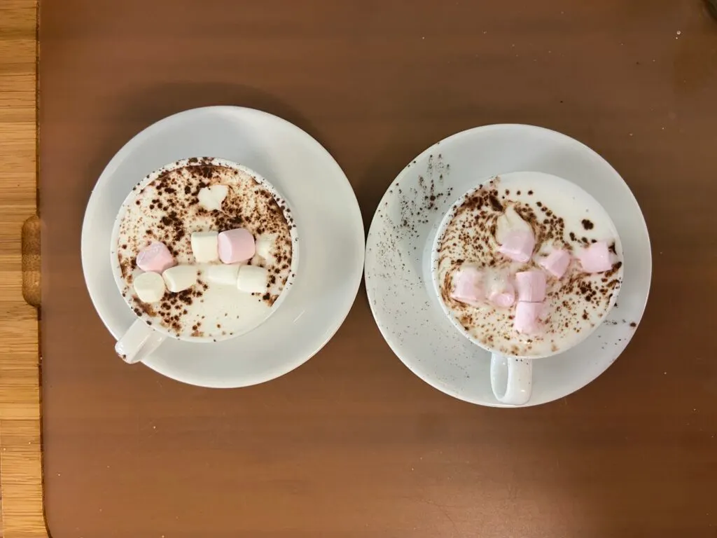 Two mugs of hot chocolate topped with cocoa powder and marshmallows on a brown surface.