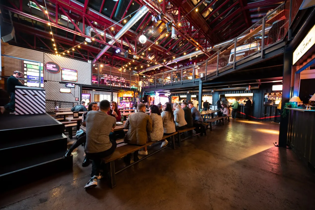 Groups of people sitting at long benches inside a modern bowling alley with neon lighting and exposed industrial ceiling.