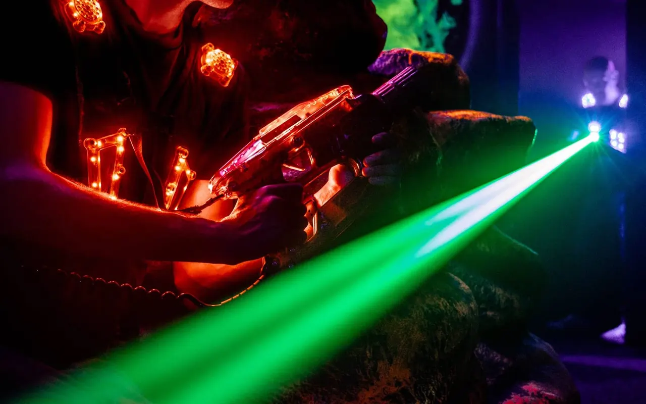 Two players aiming neon laser guns across a dark arena illuminated by colourful red and green laser beams.