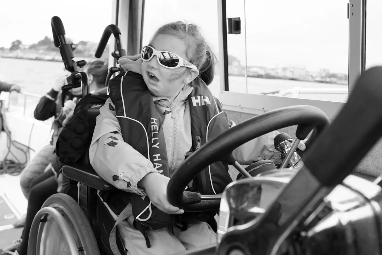 Young child at the helm of a speedboat, wearing sunglasses and a life jacket, with water visible in the background.