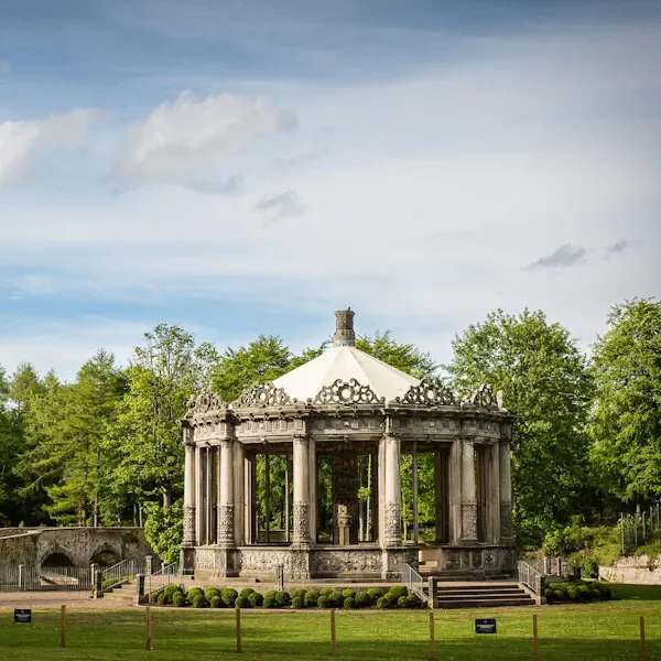 Classical stone pavilion with columns set in a manicured garden surrounded by woodland.