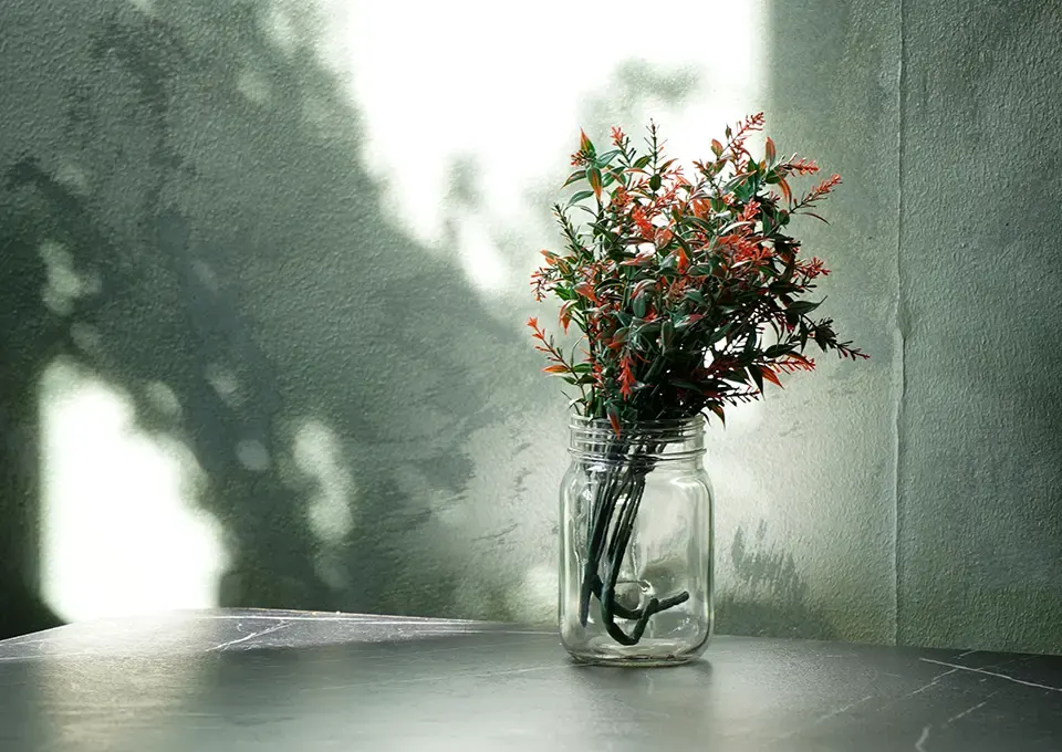 A glass jar with orange and green flowering stems sits on a table against a textured grey wall with dappled shadow.