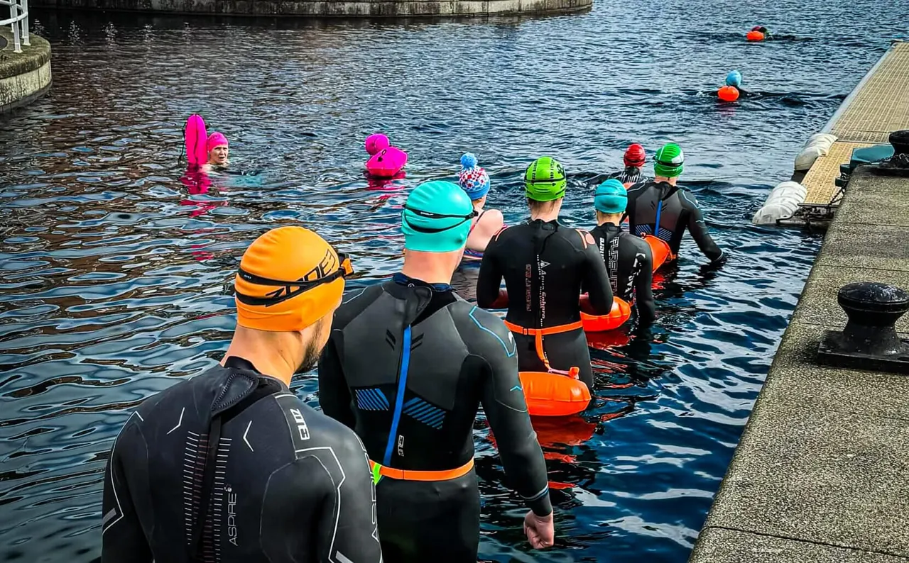 Group of swimmers in wetsuits and colourful swimming caps entering the water from a dock for a supervised swimming session.