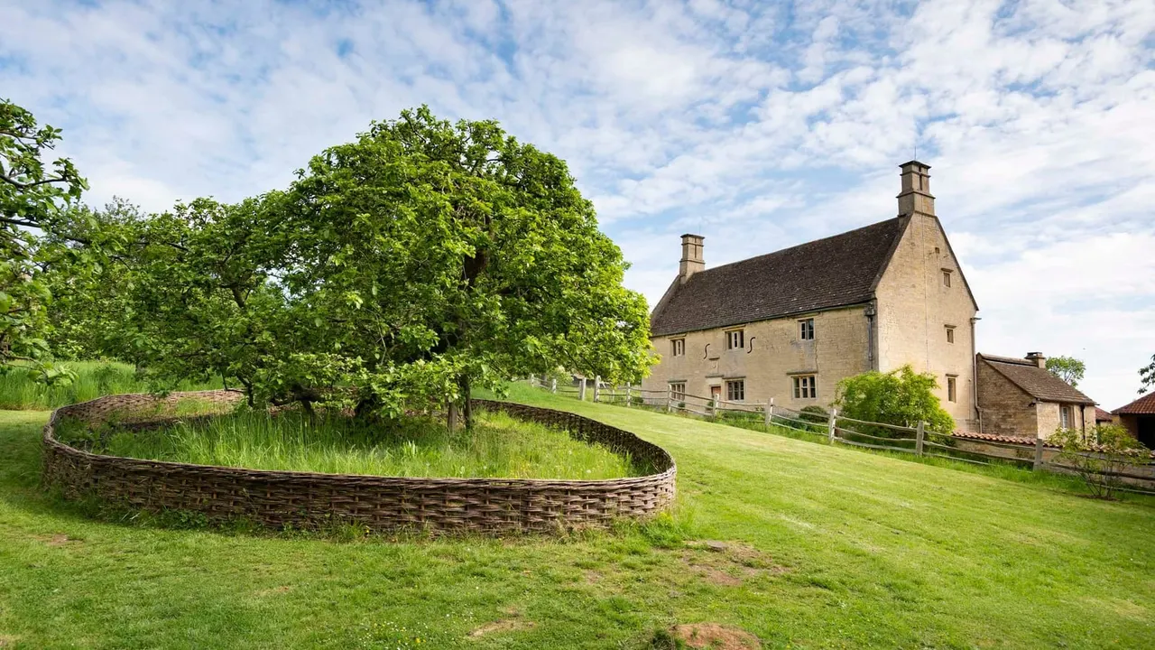 Stone farmhouse with chimneys set in green fields, with mature trees and woven fencing in foreground.