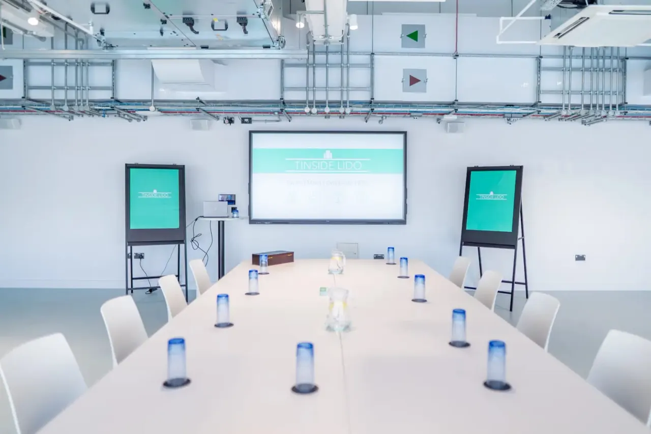 Modern meeting room with long white table, chairs, water glasses, and display screens showing Tinside Lido branding.