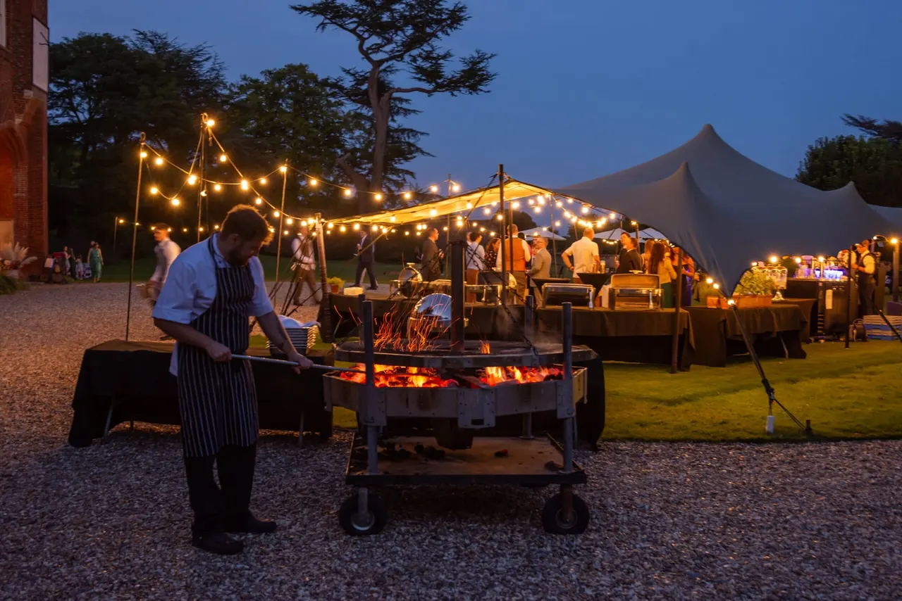 Chef cooking at an outdoor evening event with marquees, string lights, and guests gathered on the lawn at dusk.