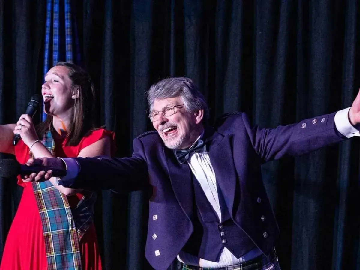 Two performers on stage singing, one in a red dress with tartan details holding a microphone, the other in formal navy jacket and tartan trousers, both smiling against a dark curtain backdrop.
