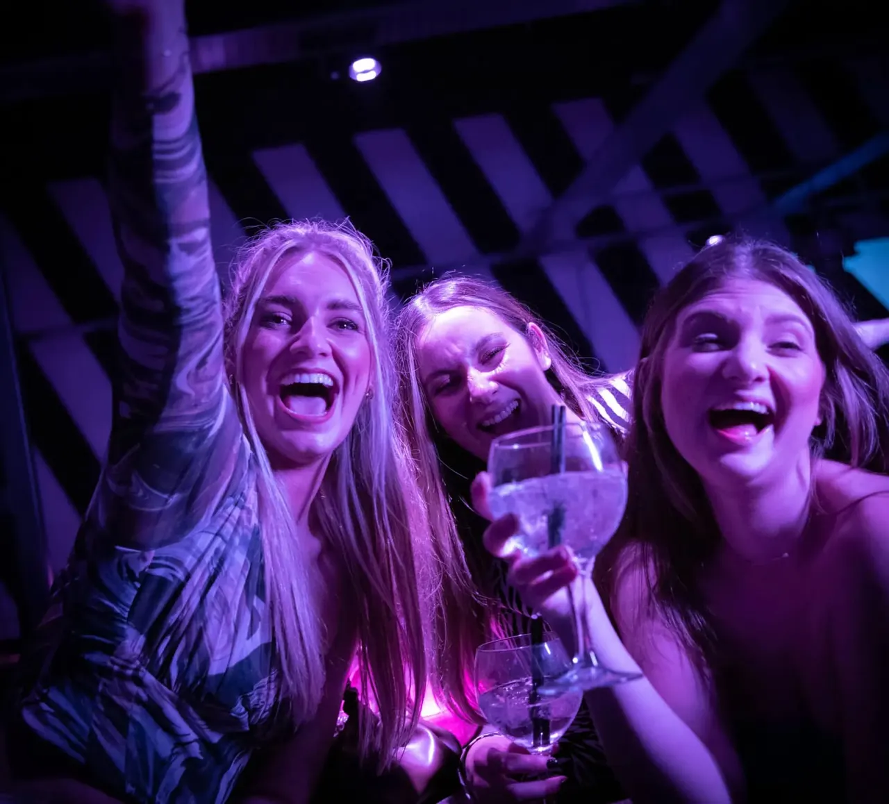 Three young women laughing and holding drinks in a vibrant purple-lit nightclub venue.