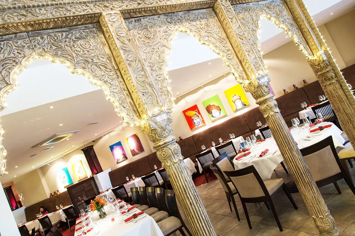 Ornate gold-framed archways frame an elegant restaurant dining room with white tablecloths and colourful artwork on walls.