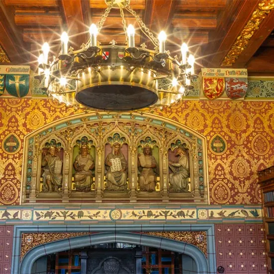 Ornate interior of a historic castle hall featuring a brass chandelier, gilt-framed religious figures in alcoves, and decorative gold patterned wallpaper.