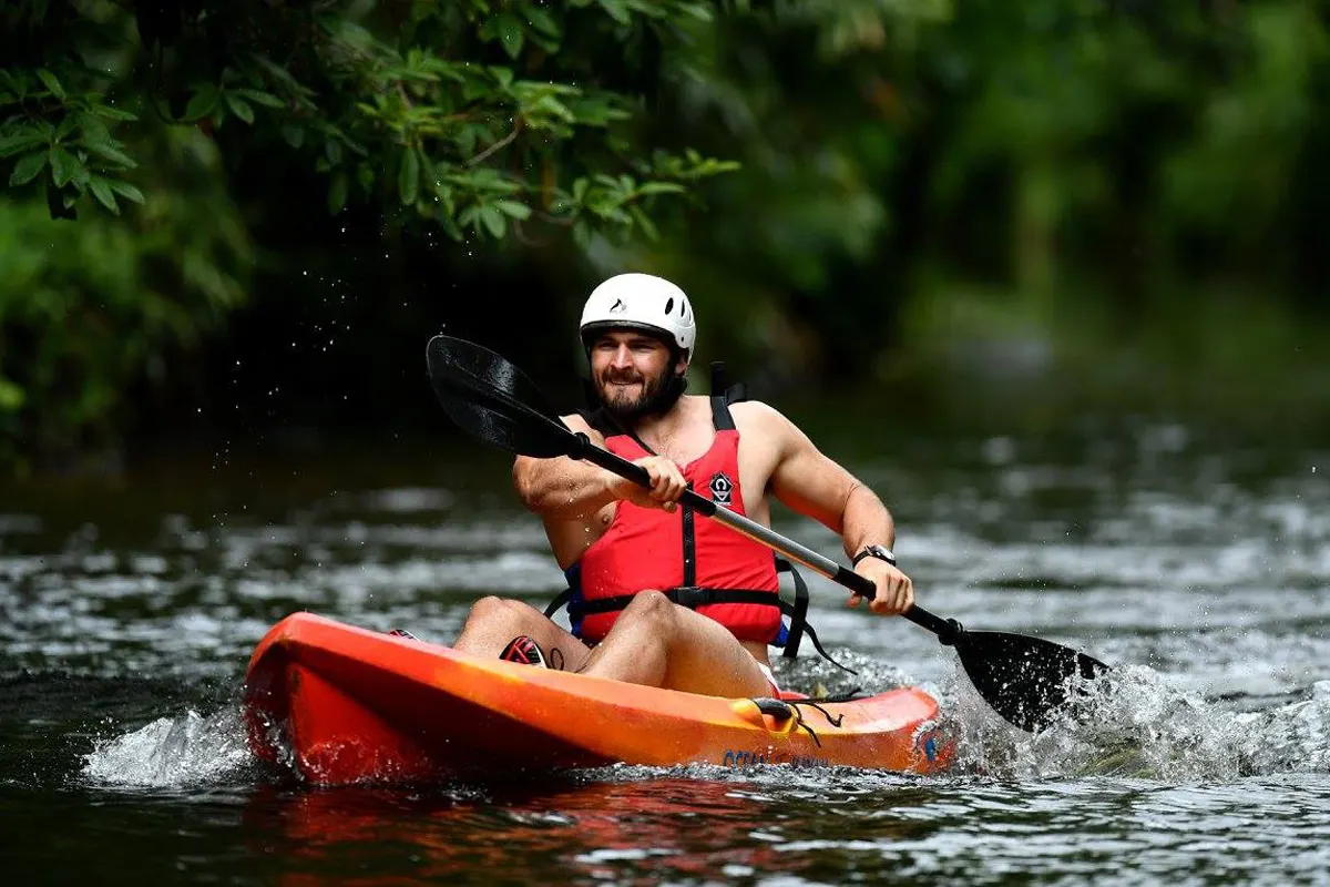 Man in white helmet and red life jacket kayaking on a river lined with green foliage.