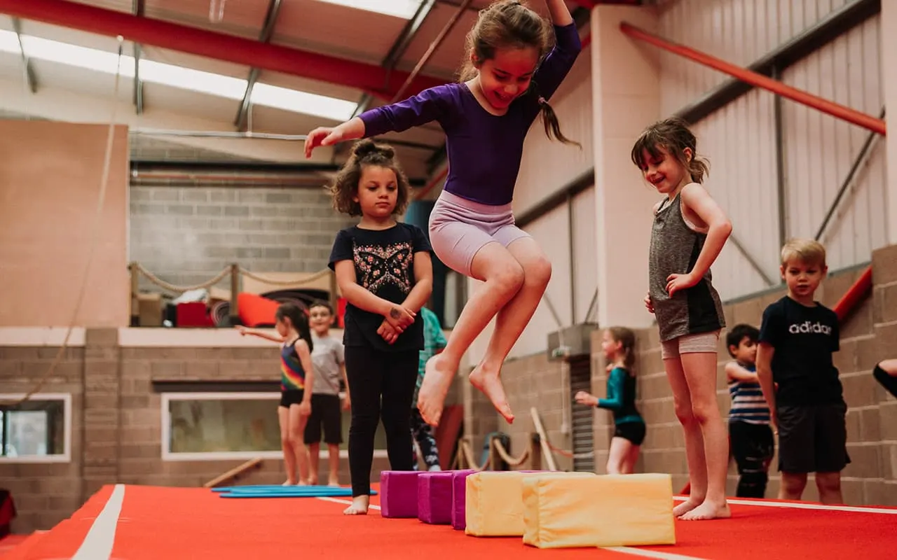 Child jumping over foam blocks on red mat during gymnastics class with other children watching.