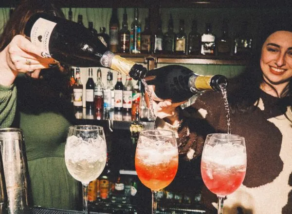 Two bartenders pouring champagne and mixing cocktails at a bar counter with bottles displayed behind.