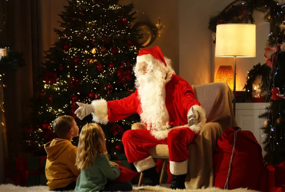 Father Christmas in red suit sits with two young children in a festive lounge decorated with illuminated Christmas tree and garlands.