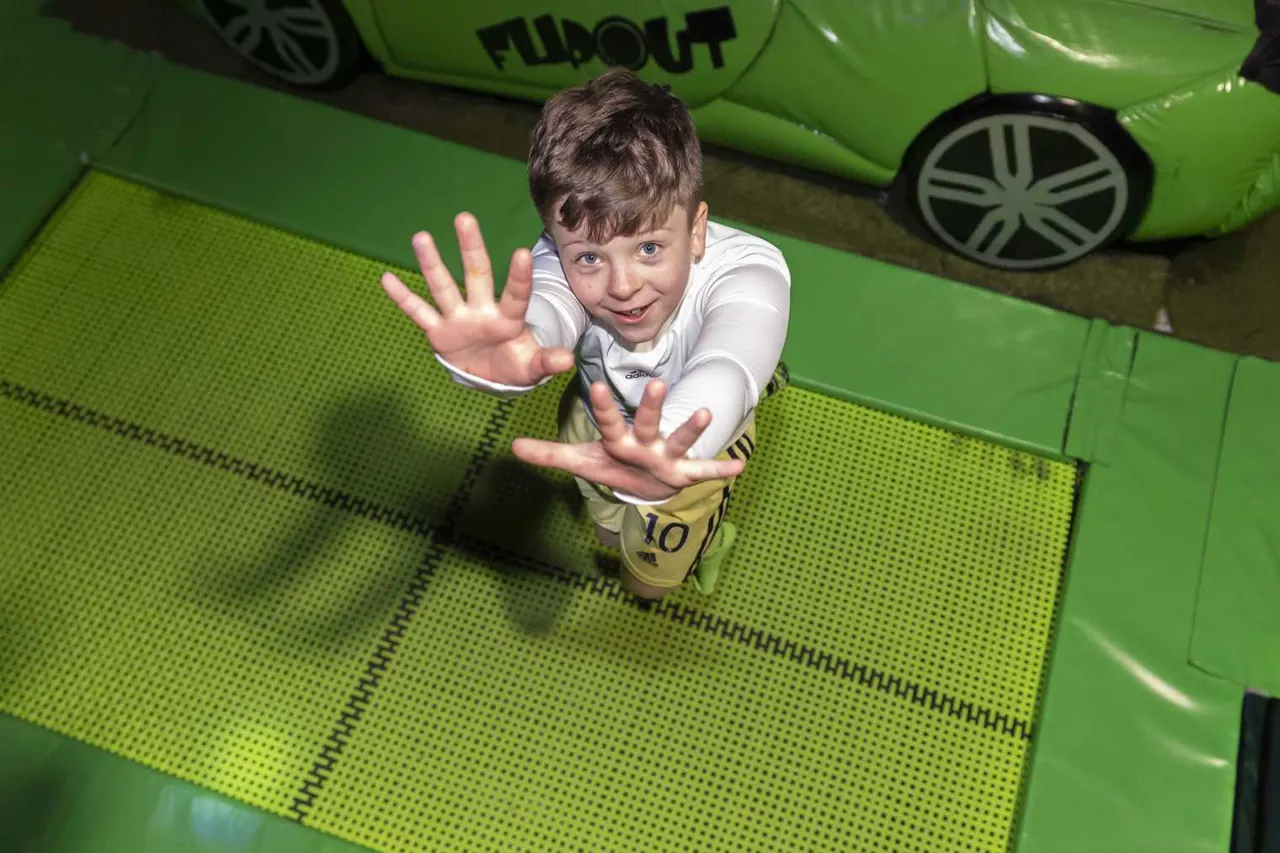 Young boy on a bright green trampoline, waving at the camera with both hands and smiling.