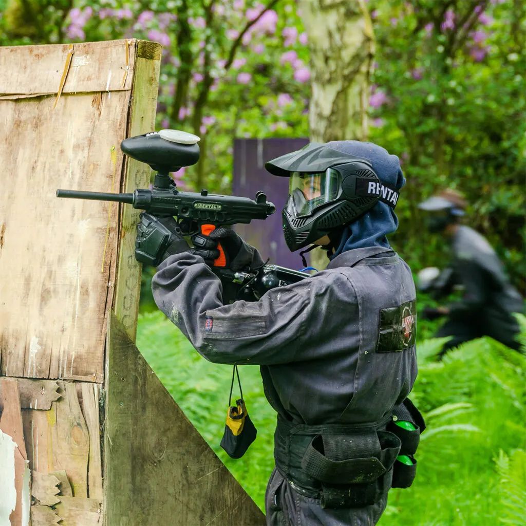 Paintball player in protective gear and mask aims paintball gun at wooden obstacle in garden setting with flowering vines.