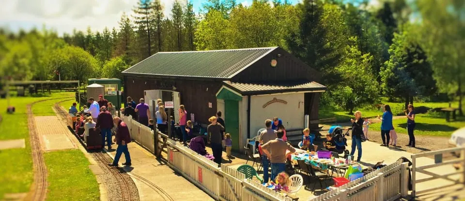Visitors gather around a miniature railway station with farm buildings in a tree-lined rural setting.