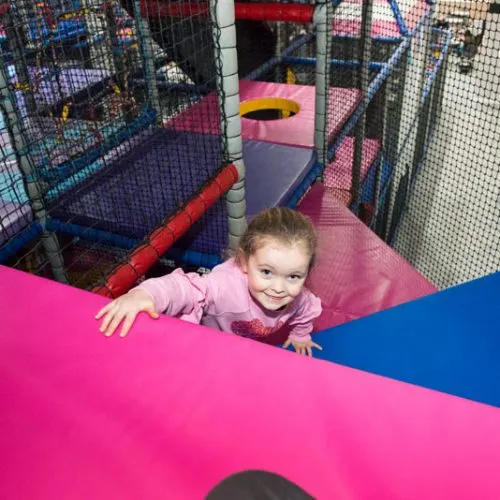 Young child playing in a colourful soft play facility with pink and blue foam structures and netting.