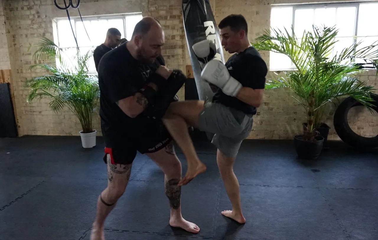 Two men practise boxing pad work in an industrial gym space with exposed brick and potted plants.