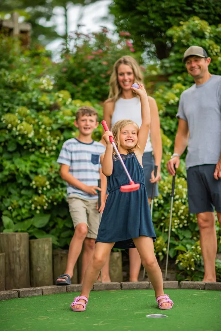 Family playing mini golf in a lush green garden setting with shrubs and flowers in the background.