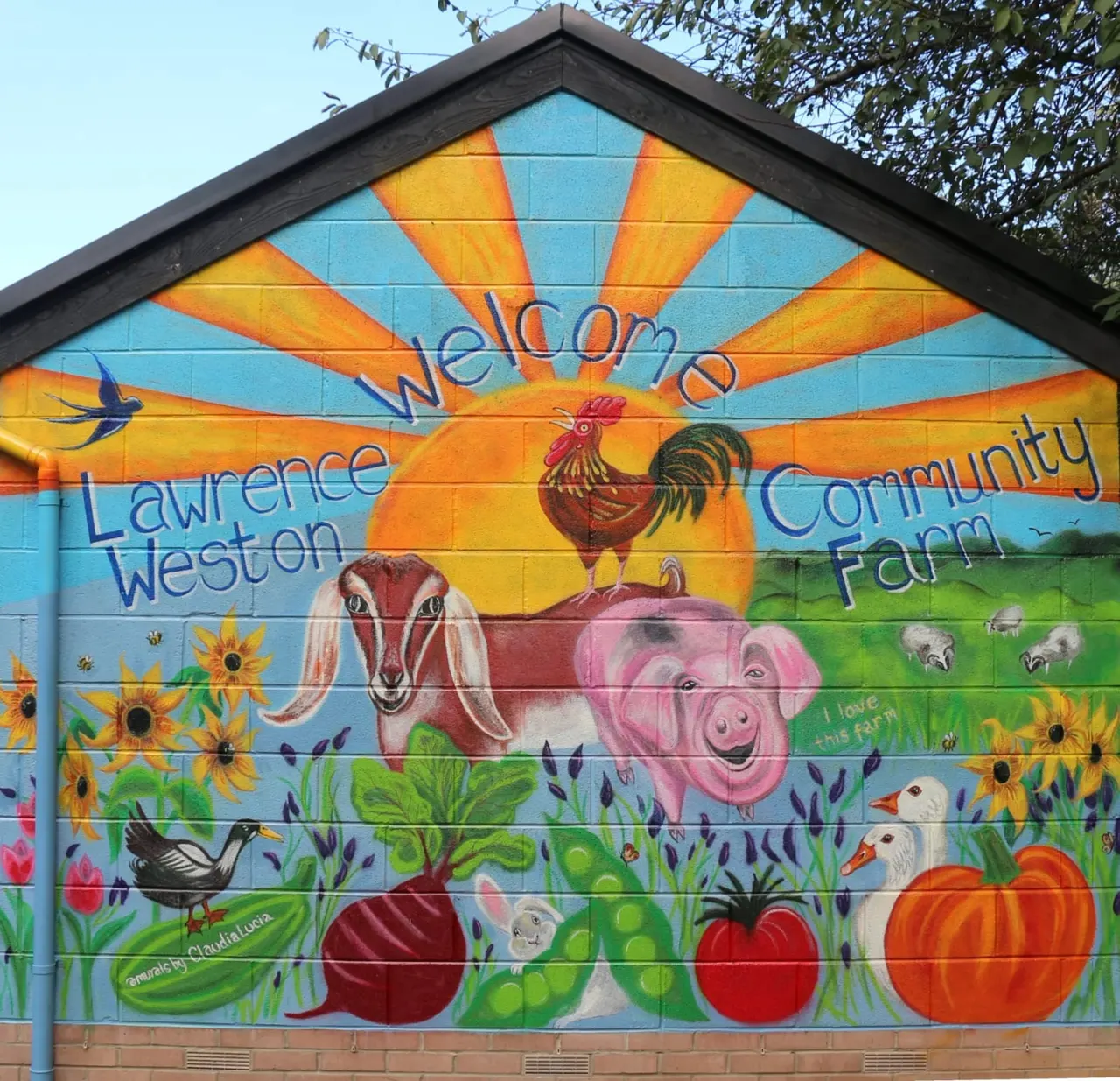 Colourful mural on farm building depicting sun, farm animals including a rooster and pig, sunflowers, vegetables and birds.