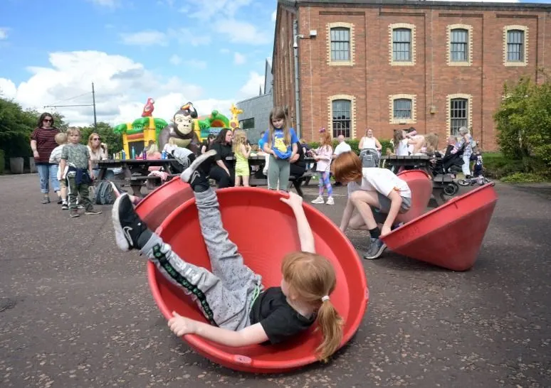 Children playing in red boat-shaped play equipment at an outdoor venue with a brick building and other visitors in the background.